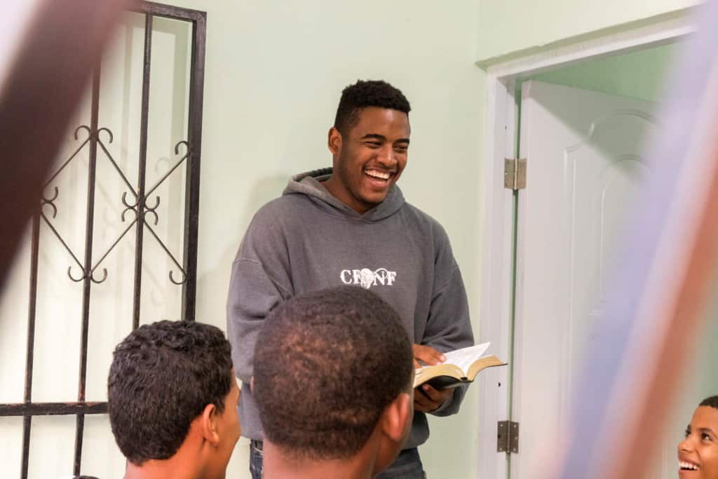 Oscar is inspiring people by speaking about identity to a group of 15-18 years old with his Bible in hand, wearing a gray sweatshirt.