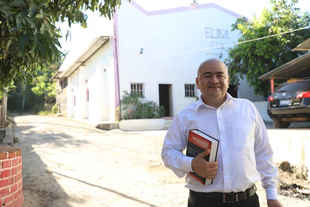 Pastor Pedro Segovia is standing by a dirt alley. He has a Bible in his hand. He is wearing a white button-down shirt and black pants. The background is the Church’s temple with the word ‘ELIM’ which is the name of the evangelical denomination. Pastor Pedro is smiling and making eye contact with the camera.