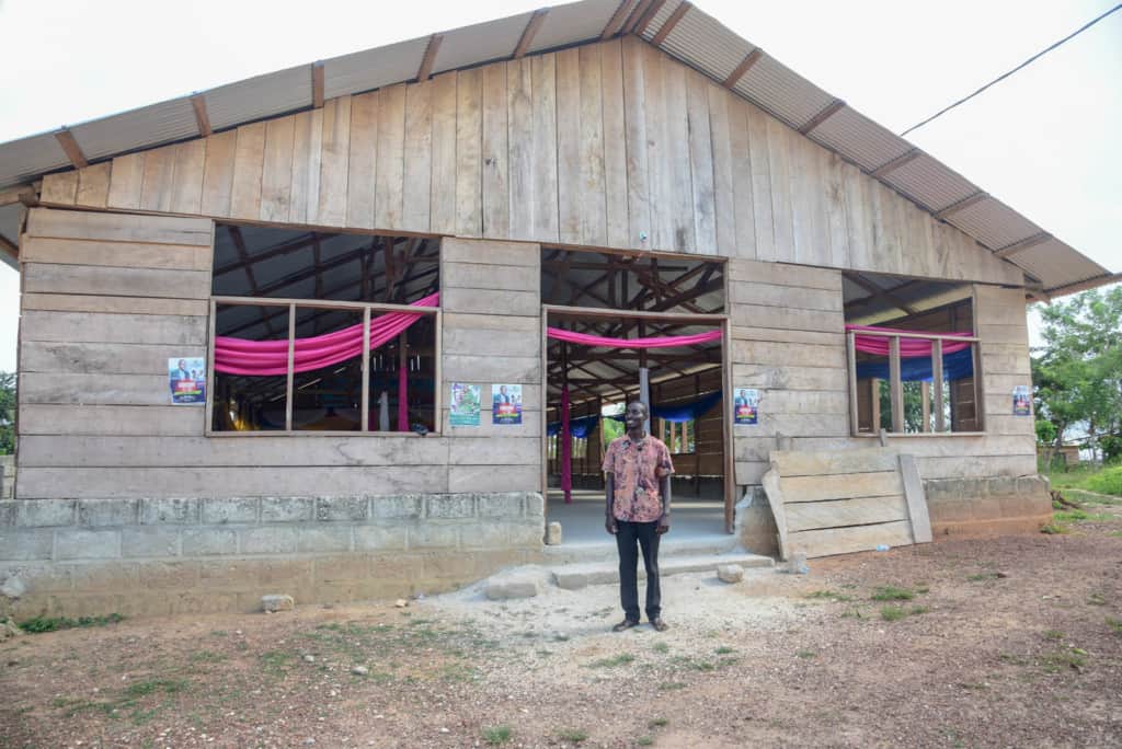 Pastor Joseph is standing in front of a new building in this photo of churches around the world. He is wearing dark pants and a pink patterned shirt.