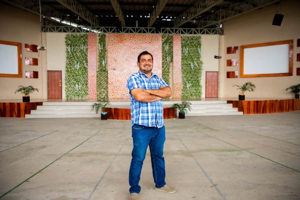 Fabricio is wearing a blue and white plaid shirt and jeans. He is standing at the church with his arms crossed. The pulpit is in the background.