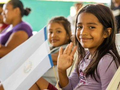 A girl in a purple shirt is sitting in a chair, smiling and waving. She is holding a white paper flag on a stick.