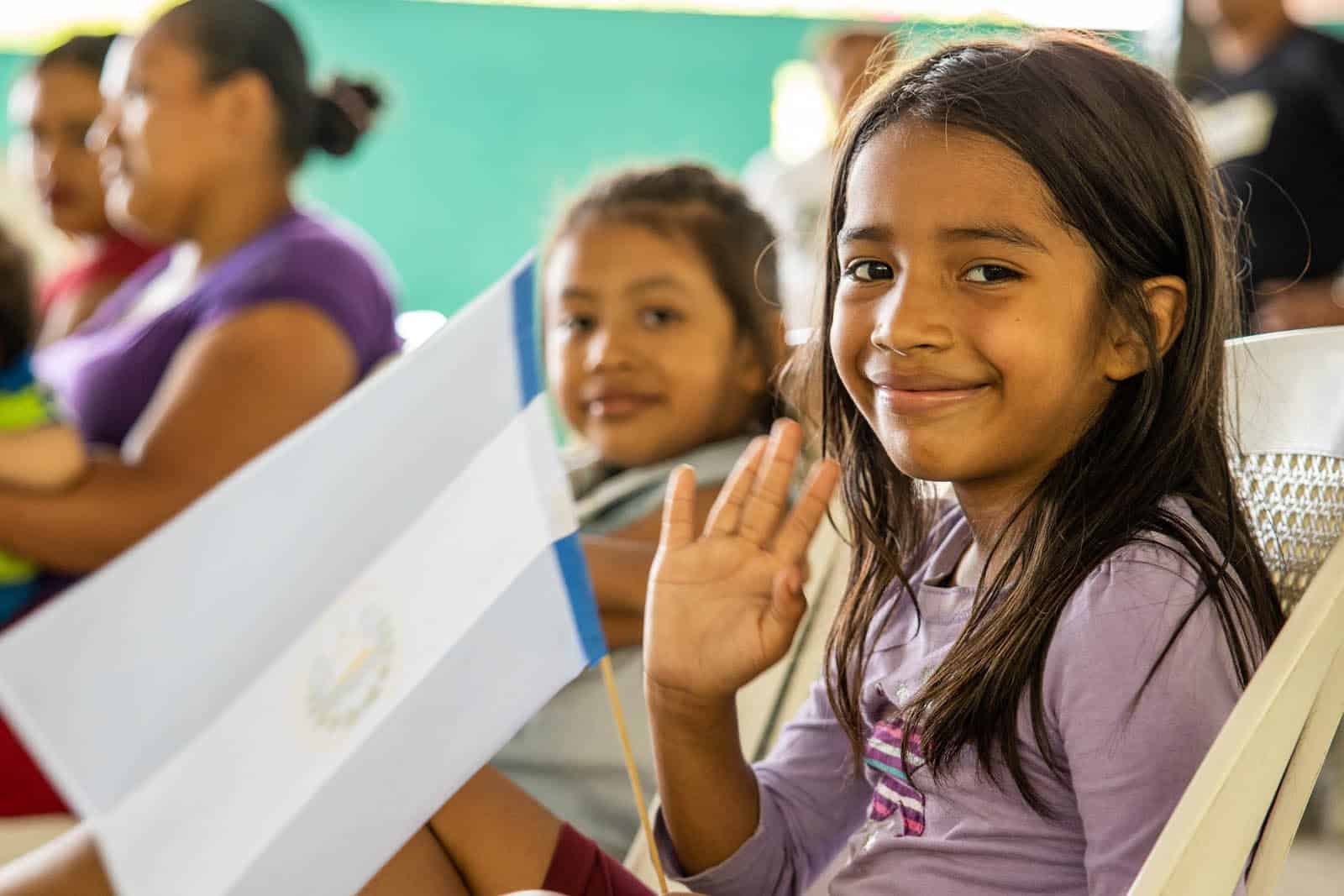 A girl in a purple shirt is sitting in a chair, smiling and waving. She is holding a white paper flag on a stick.