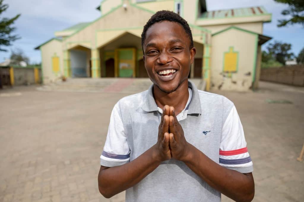 Kelvin is wearing a white shirt and jeans. He is standing outside the Compassion center and church with his hands touching in front of him. The building is yellow and green.