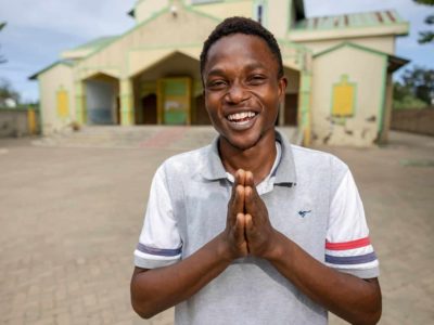 Kelvin is wearing a white shirt and jeans. He is standing outside the Compassion center with his hands touching in front of him. The Compassion center building is yellow and green.