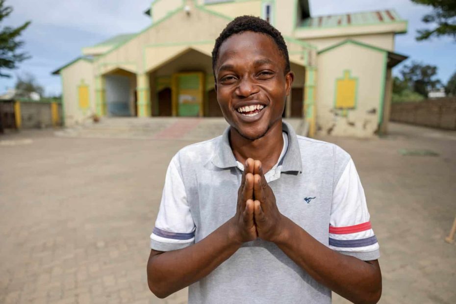 Kelvin is wearing a white shirt and jeans. He is standing outside the Compassion center with his hands touching in front of him. The Compassion center building is yellow and green.