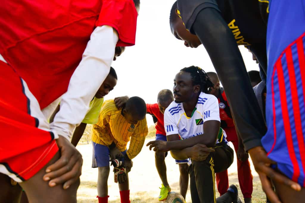 Amani, one of our inspiring people, is seen here kneeling down, wearing a white and blue shirt. The children are standing all around him while he talks to them.