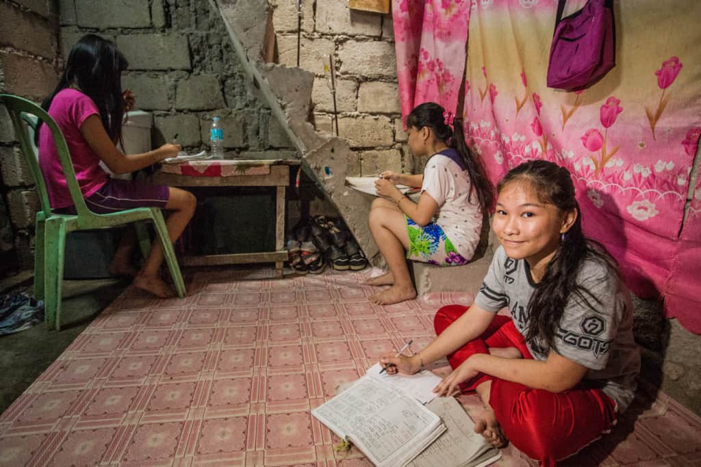 Two of the sisters are sitting on the floor working, and one sister is sitting in a green chair at a desk. There is a pink and yellow sheet hanging on the wall.