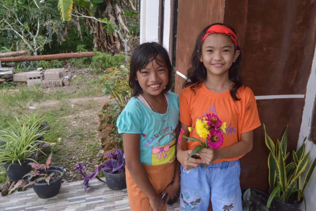 Keysha helps others. She is wearing an orange shirt and light jeans. She is standing outside with her friend and is holding a bouquet of pink and yellow flowers.