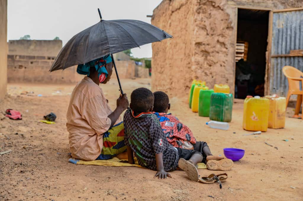 Alima is wearing a pink shirt and a colorfully patterned skirt. She is holding up a black umbrella. Her two grandsons, Fatafou and Kassoum, are sitting with her. In front of their home are plastic chairs and jerry cans.