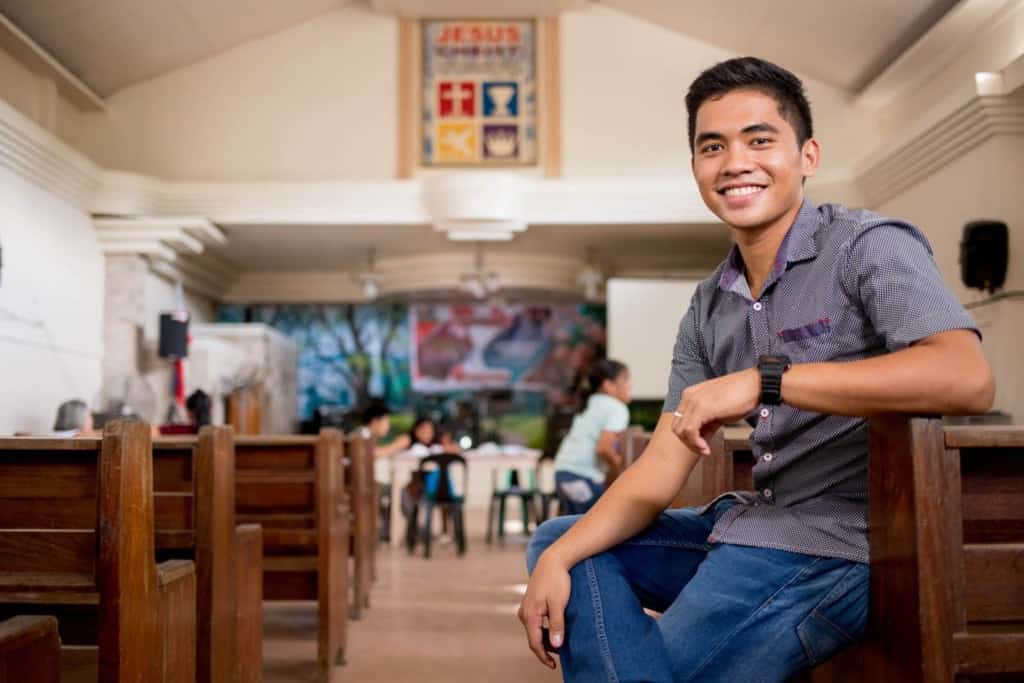 Stephen, in a gray shirt and jeans, is at the center sitting on the arm of one of the pews as he poses for a picture.