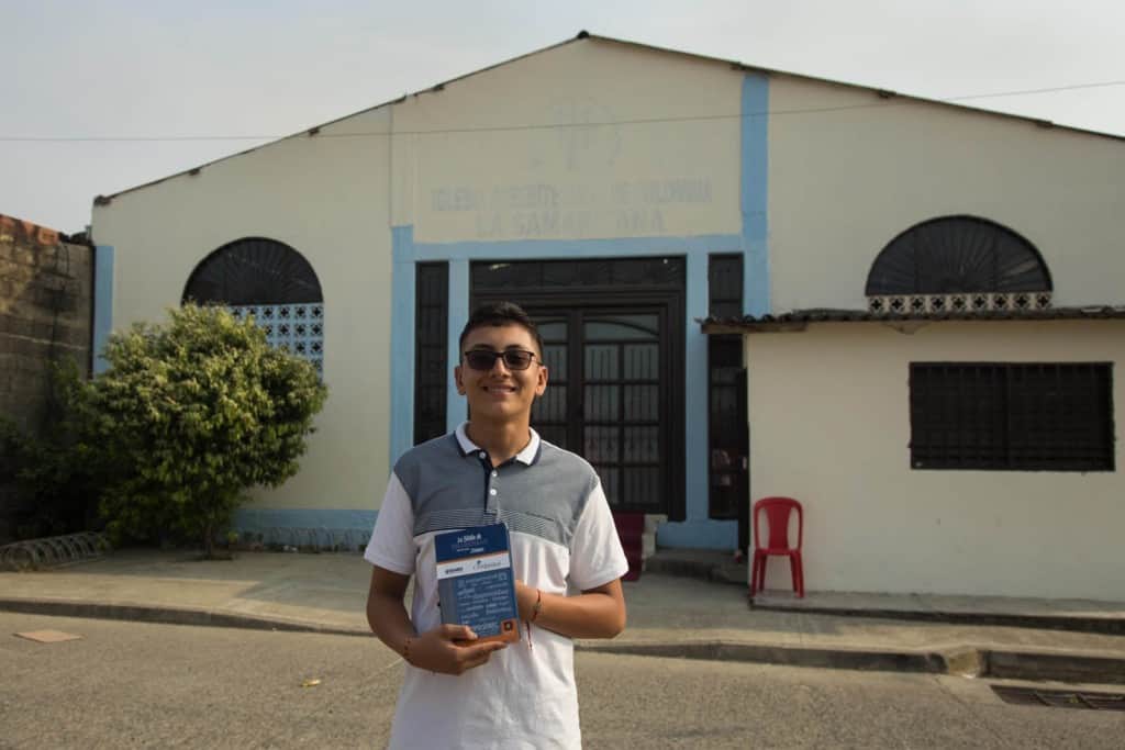 Julian, in a gray and white shirt and sun glasses, is holding the blue Bible he received at the project during a special event. He is standing in front of the church, which is pale yellow and has a black door and blue trim.