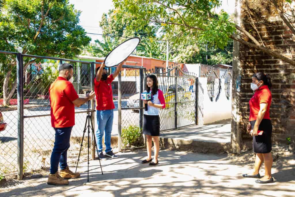 Cristina is wearing a white shirt with pink sleeves and a black skirt. She is being recorded with a cell phone in front of her Compassion center, sharing a word of encouragement.