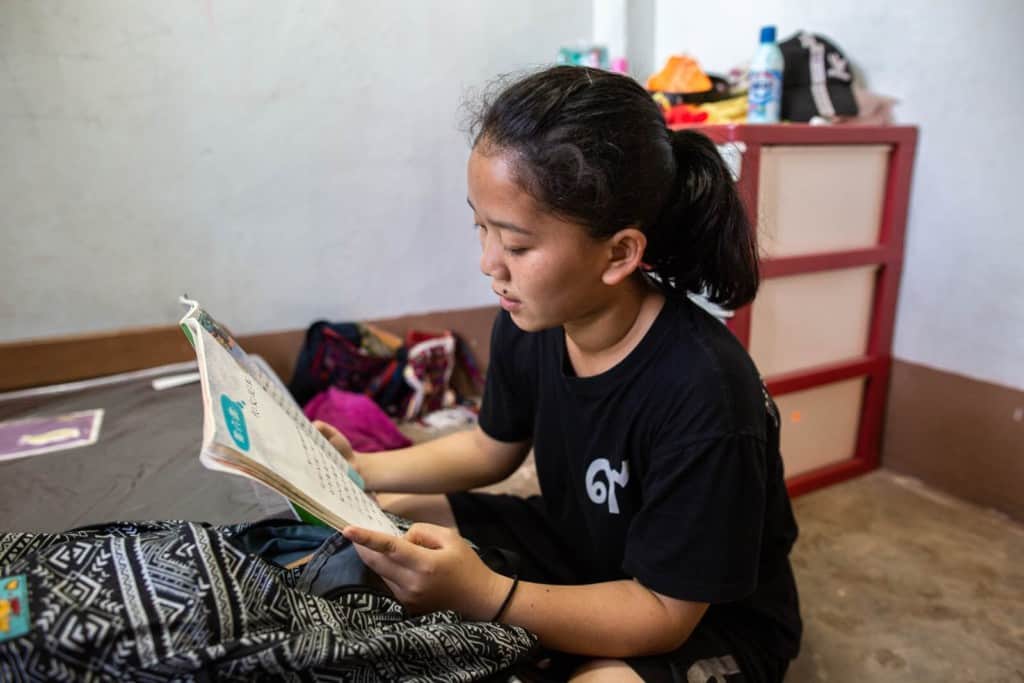 Chanikan is sitting in her bedroom and reading her Chinese homework. She is wearing a black tee shirt and is reading a book.
