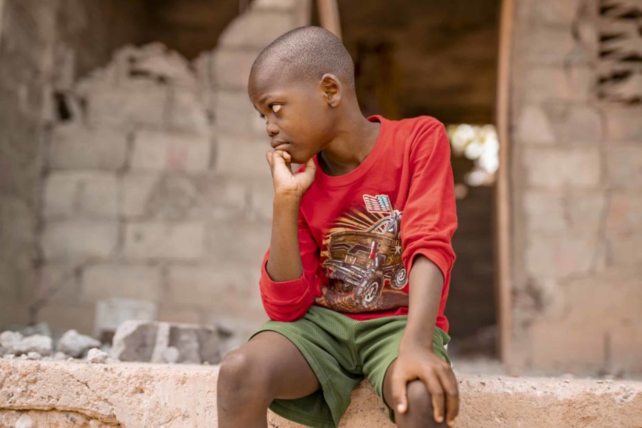 A Haitian boy wearing a red shirt and green shorts has a somber expression. He is sitting in front of a building damaged in a 2018 earthquake in Haiti.