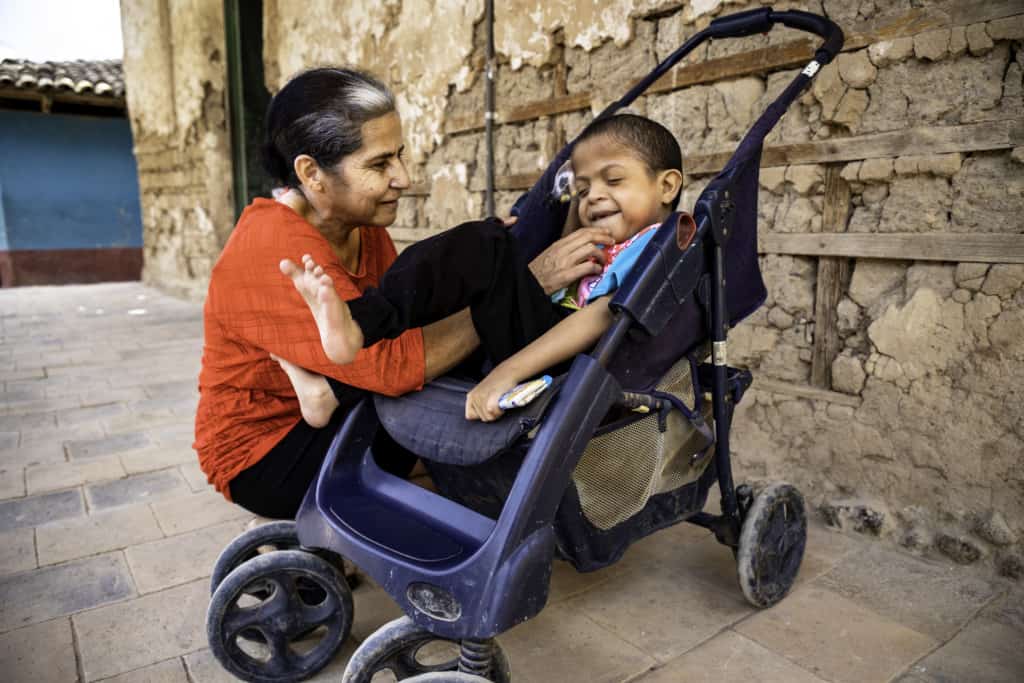 A mother smiles and plays with her son, who is smiling in a wheelchair