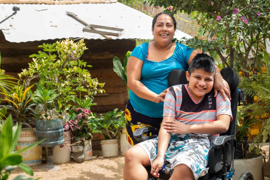 A boy smiles as he sits in a wheelchair. His mother stands behind him with her arms on his shoulders.