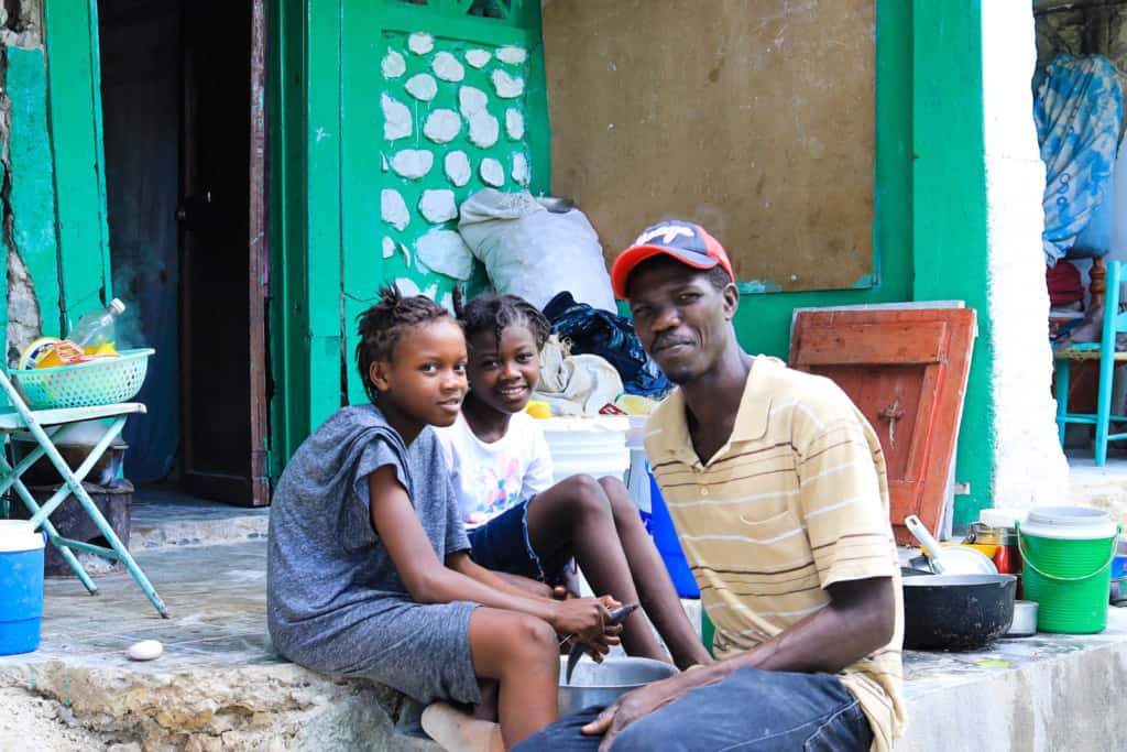 Samantha, wearing a gray shirt and gray shorts, is sitting outside her home with Sandra, wearing a white shirt and dark shorts. Their father, Sony, is next to them. Their house was damaged in the earthquake.