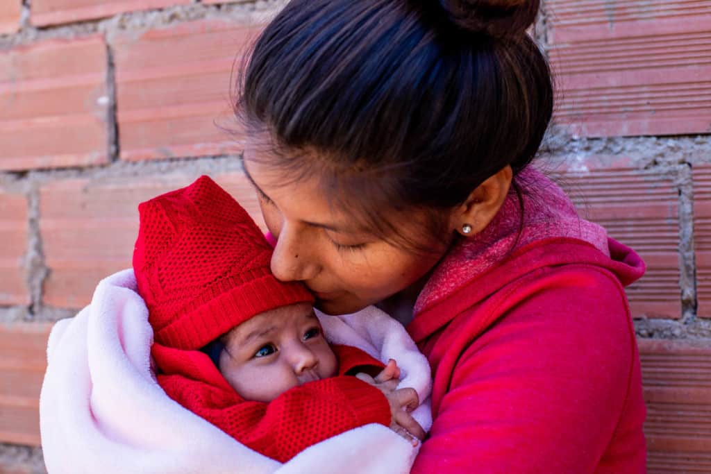 Saida is wearing a red sweatshirt and black leggings. She is holding her baby, Salome, wearing a red outfit and is wrapped in a pink blanket. They are standing in front of a brick wall.