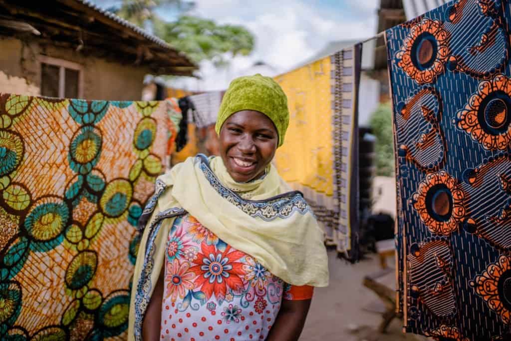 Warda, in a yellow scarf and green hat, is standing in front of laundry hanging on clotheslines outside her home.