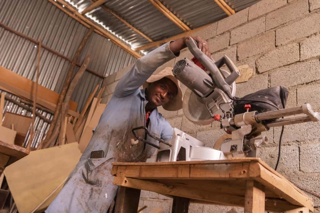 Sintayehu uses a saw in his carpentry shop. He is wearing a long blue coat and a hat.