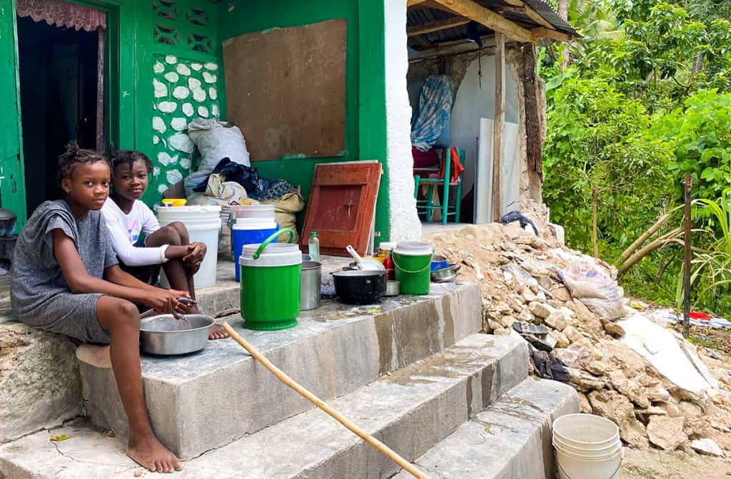 Two girls sit on a concrete porch. They are holding a fish to prepare for a meal. There is rubble from a partially collapsed home.