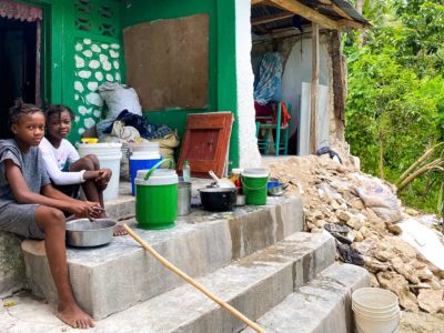 Two girls sit on concrete stairs near rubble of a home that is partially collapsed. They are preparing a fish to eat.