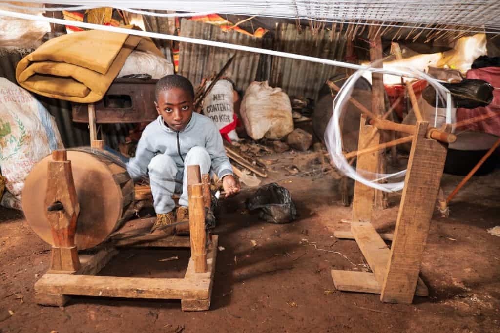 A boy wearing boots and a light colored outfit sits near a cotton wheel in Ethiopia. He is helping his father make fabric to sell at a market.