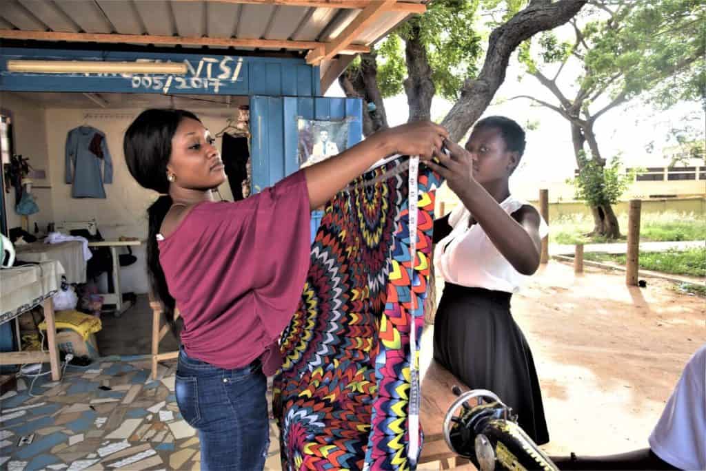 Two women in Ghana hold opposite sides of a colorful piece of fabric. One is holding a measuring tape to the fabric. She is wearing a pink shirt and blue jeans.
