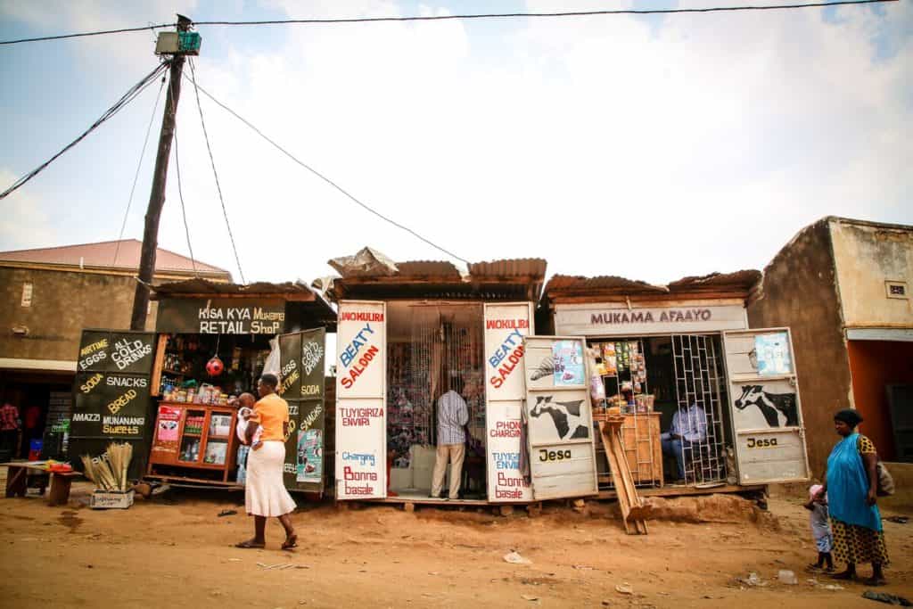 Shops line a dirt road in Uganda. Women and children walk past.