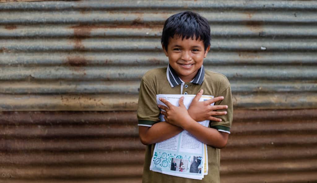 Weslin is wearing a brown shirt with jeans. He is standing in front of his home and is holding some of his sponsor's letters close to his chest. His home is made of corrugated metal sheets.