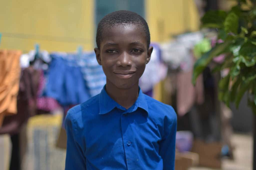 Justice is wearing dark blue shorts and a dark blue shirt. He is standing outside his home and there are clothes hanging on a clothesline behind him.