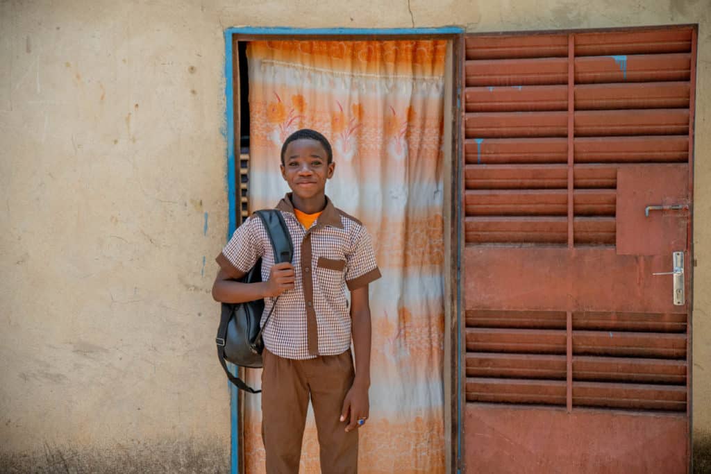 Sansan is wearing a brown and white shirt with tan pants. He is standing outside his home and is holding his back pack. Behind him there is an orange and white curtain on his door.