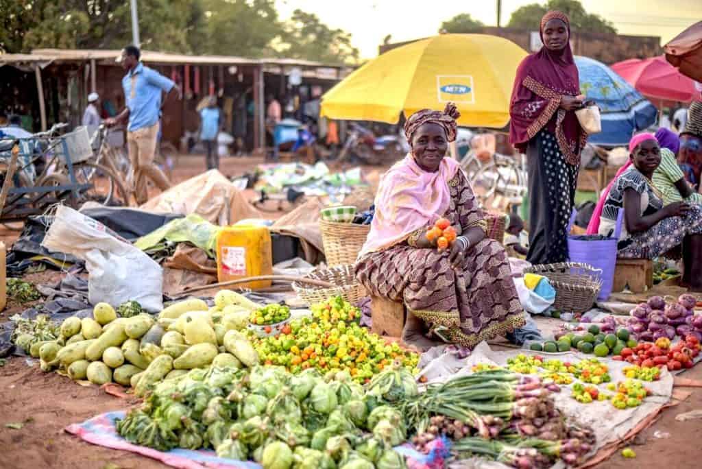 A smiling woman sits near blankets on the ground, which are covered with produce for sale. The open-air market in Burkina Faso is busy with people shopping and selling.