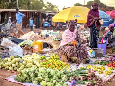 A smiling woman sits at a marketplace in Burkina Faso surrounded by fruits and vegetables on blankets.