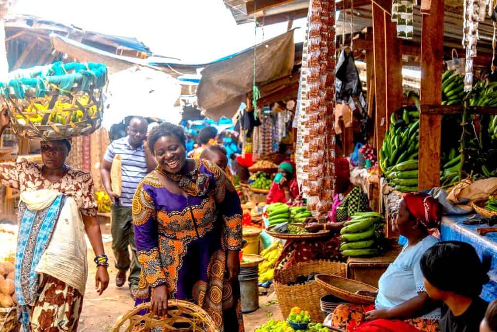 I busy outdoor marketplace in Tanzania. A smiling woman is wearing a colorful purple and orange dress. Vegetable stands selling plantains and other produce are seen behind her. A woman walks by carrying a basket of fruit on her head.