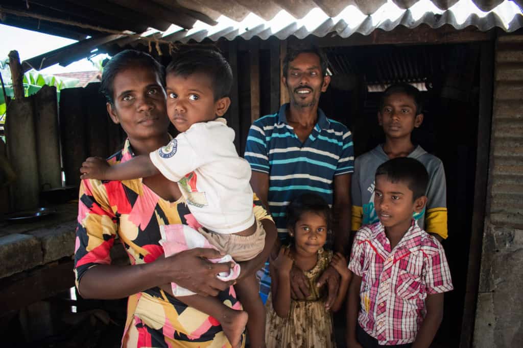 Pradeepkumar and Devaki are standing inside their home with their four children: Shalintha, Pivishan, Rekonshan, and Rebeaka. Devaki is carrying her youngest son Rekonshan. 