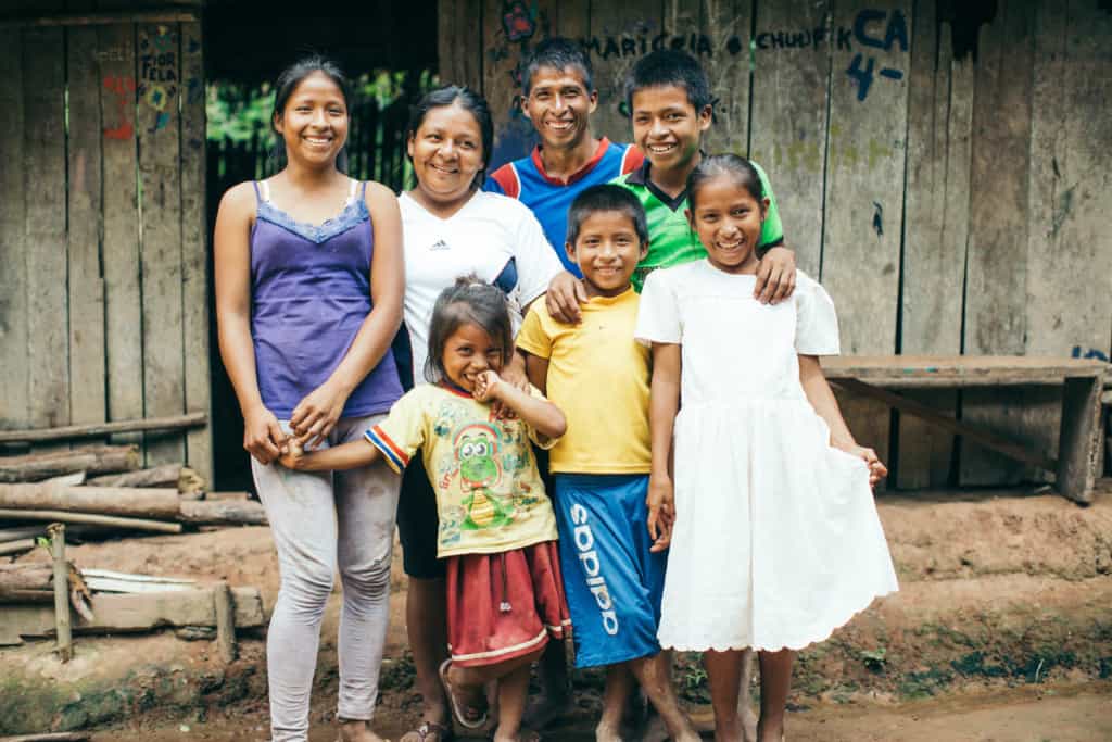 A smiling family, mother, father and children stand outside posing for a picture with their arms around each other. They wear purple, blue, white, green and yellow shirts and one girl wears a white dress. They are in front of a home made of wood boards and a thatched roof.