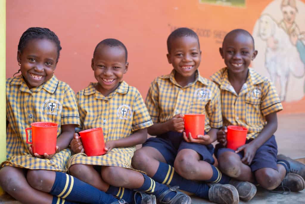 Four children in uniforms sitting on the ground. They are all holding red cups.