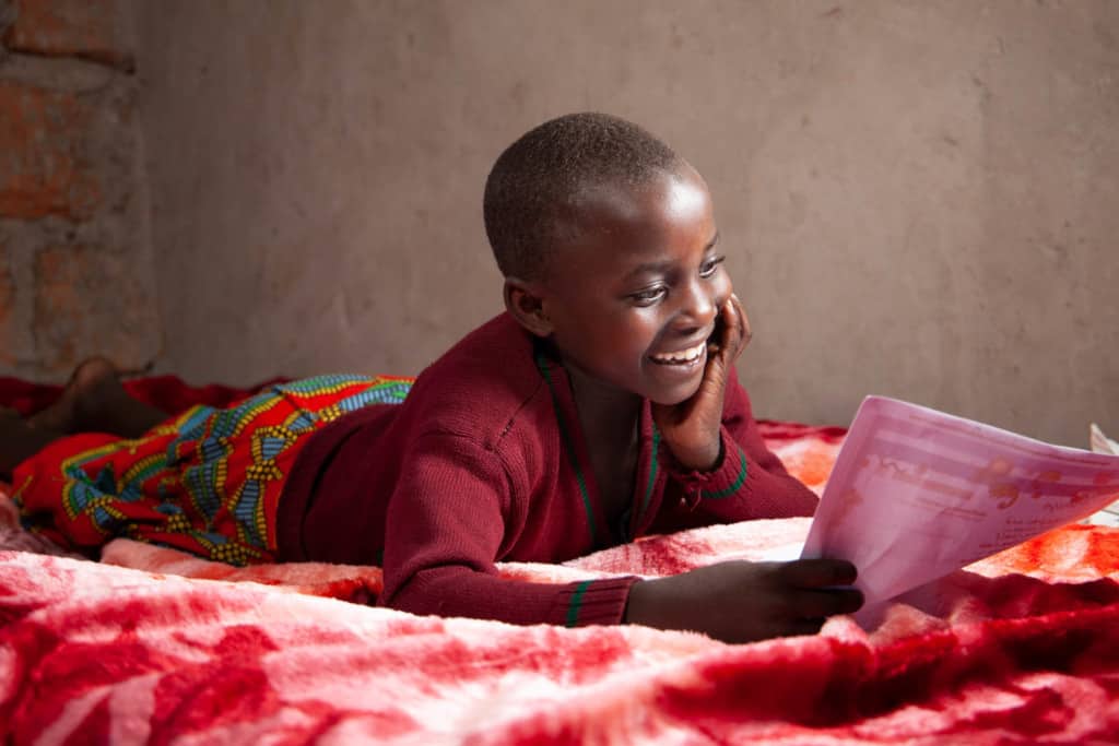 Promise is wearing a maroon shirt with a red and blue patterned skirt. She is laying on her bed and is reading a letter from her sponsor.
