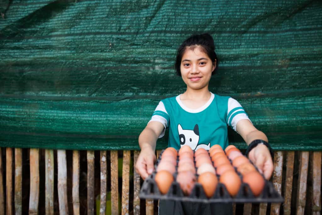 Rungnapa is showing an egg tray after she collected eggs at the agriculture project site. She is wearing a green tee shirt and is standing in front of a green tarp. Rungnapa is smiling at the camera.