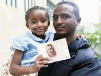 A father holds his daughter, who is holding a photo of a woman