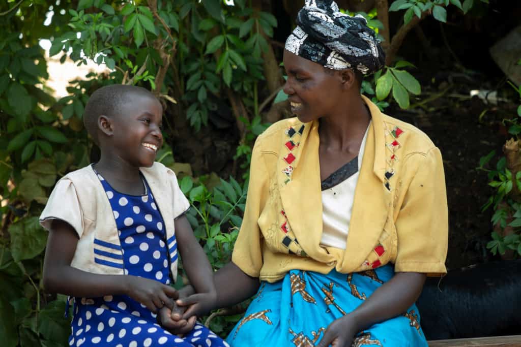 Promise is wearing a blue and white polka dotted dress with a white jacket. She is sitting outside next to her mother, Olive, wearing a yellow shirt and blue skirt. Promise is holding her mother's hand. There are trees behind them.