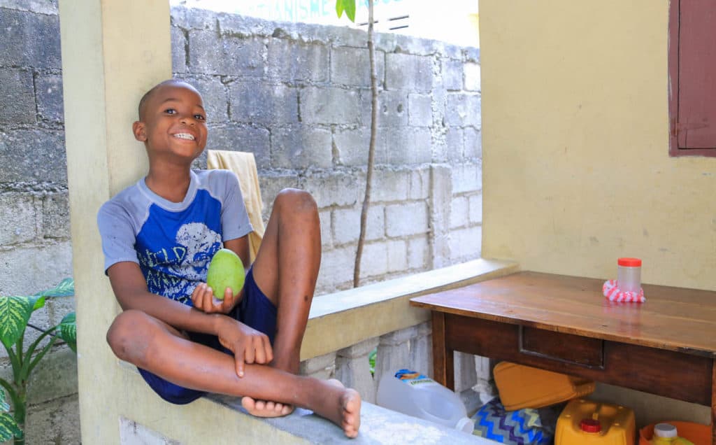 Kenderson is wearing a blue shirt and shorts. He is sitting on the front porch of his home. There is a cinder block wall behind him. He dreams of being a soccer player.