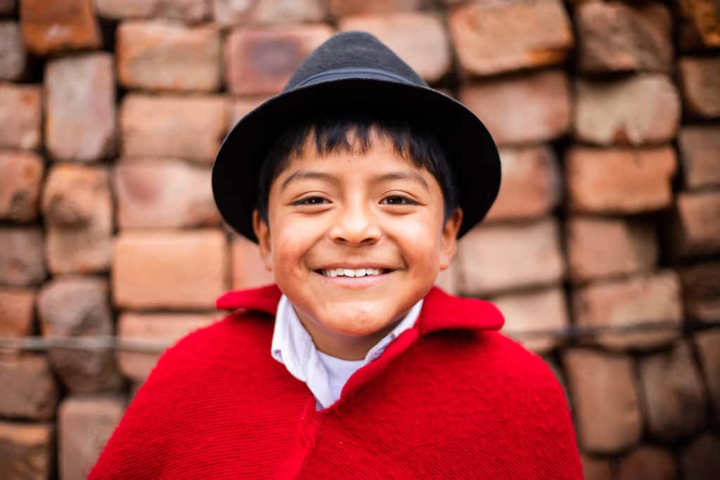 Jostin is wearing traditional clothing, a black hat and a red cape. He is standing in front of a brick wall.