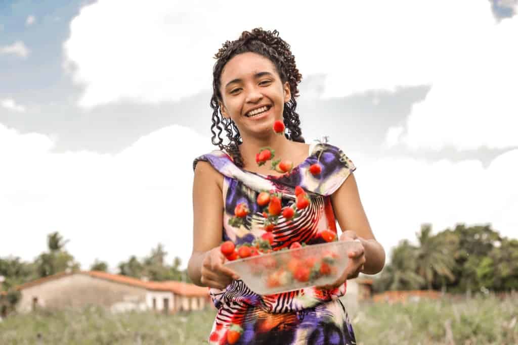 Grazielle is wearing a purple patterned dress. She is is standing outside with a plastic bowl full of strawberries that she is tossing up into the air.