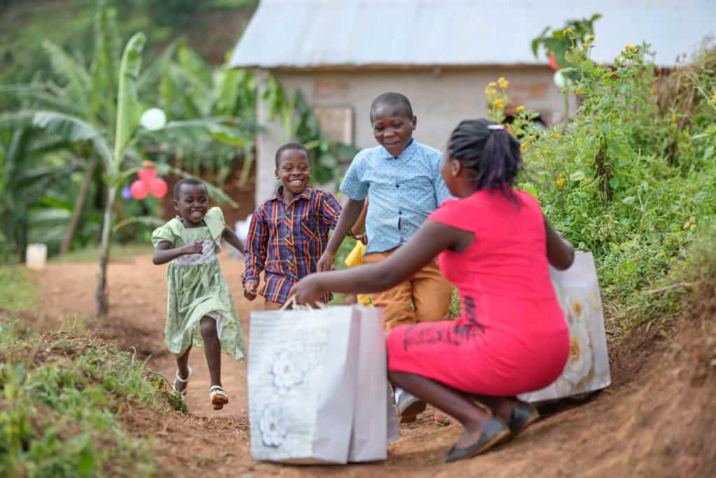 Martha is wearing a red dress and is kneeling down with bags full of Christmas gifts for the children. A large group of children is running toward her.