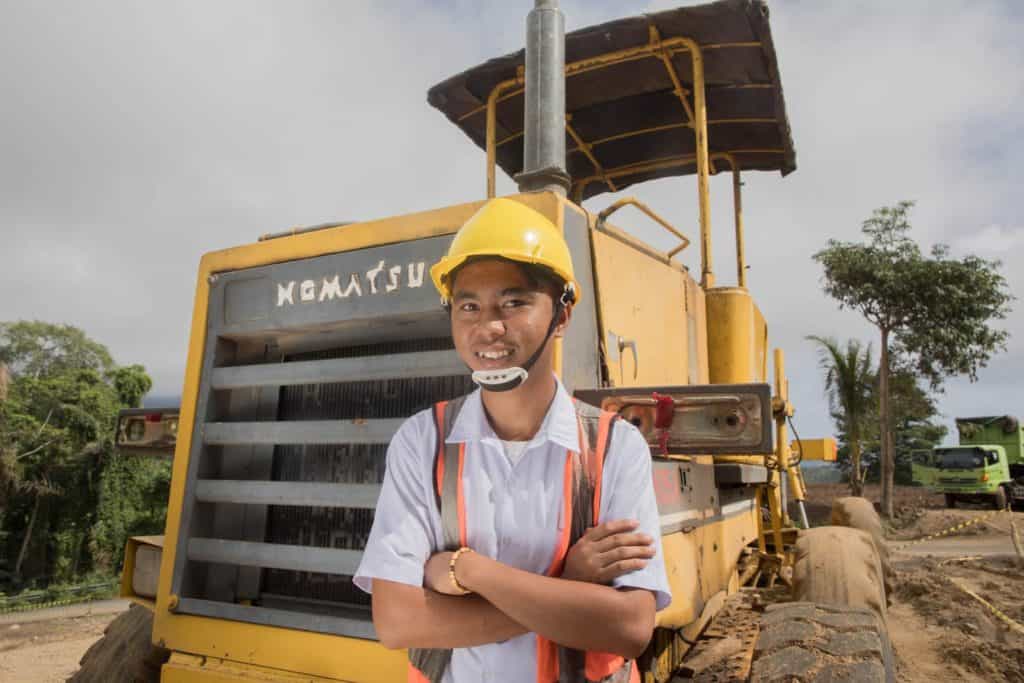 Buyung is wearing a white shirt, orange vest, and yellow hard hat. He is standing with his arms crossed in front of him. He is standing in front of a large yellow grader that is parked on the side of the road. He dreams of operating machines as a career.