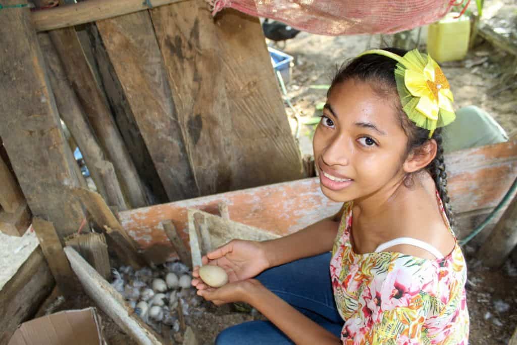 Steicy is wearing a floral shirt and a yellow bow in her hair. She is is her yard gathering some of her duck eggs. She dreams of being a graphic designer.