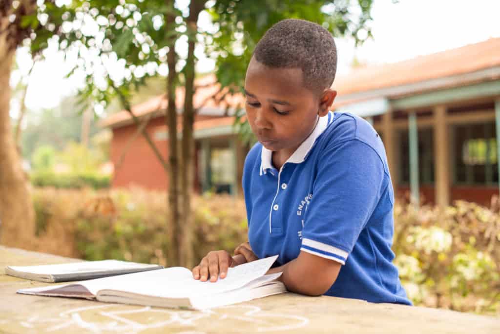 Lilian is wearing a blue polo shirt. She is sitting at a table outside her school and is reading a book.