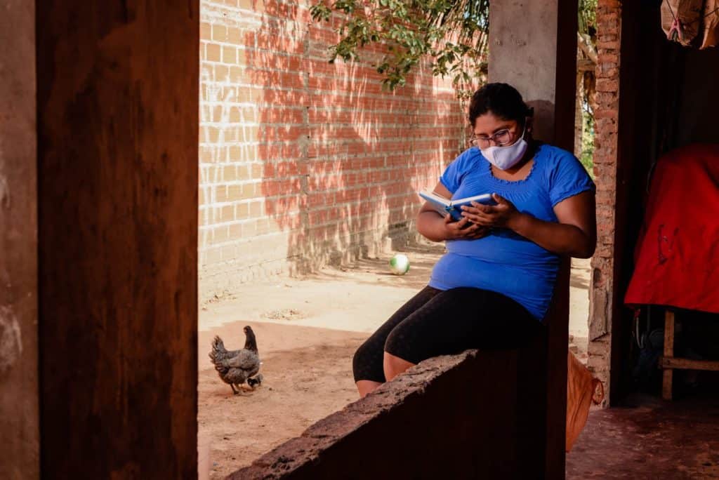 Ana is wearing a blue shirt, black shorts, and a whtie face mask. She is sitting outside her home and is reading a book. There is a chicken in the background.